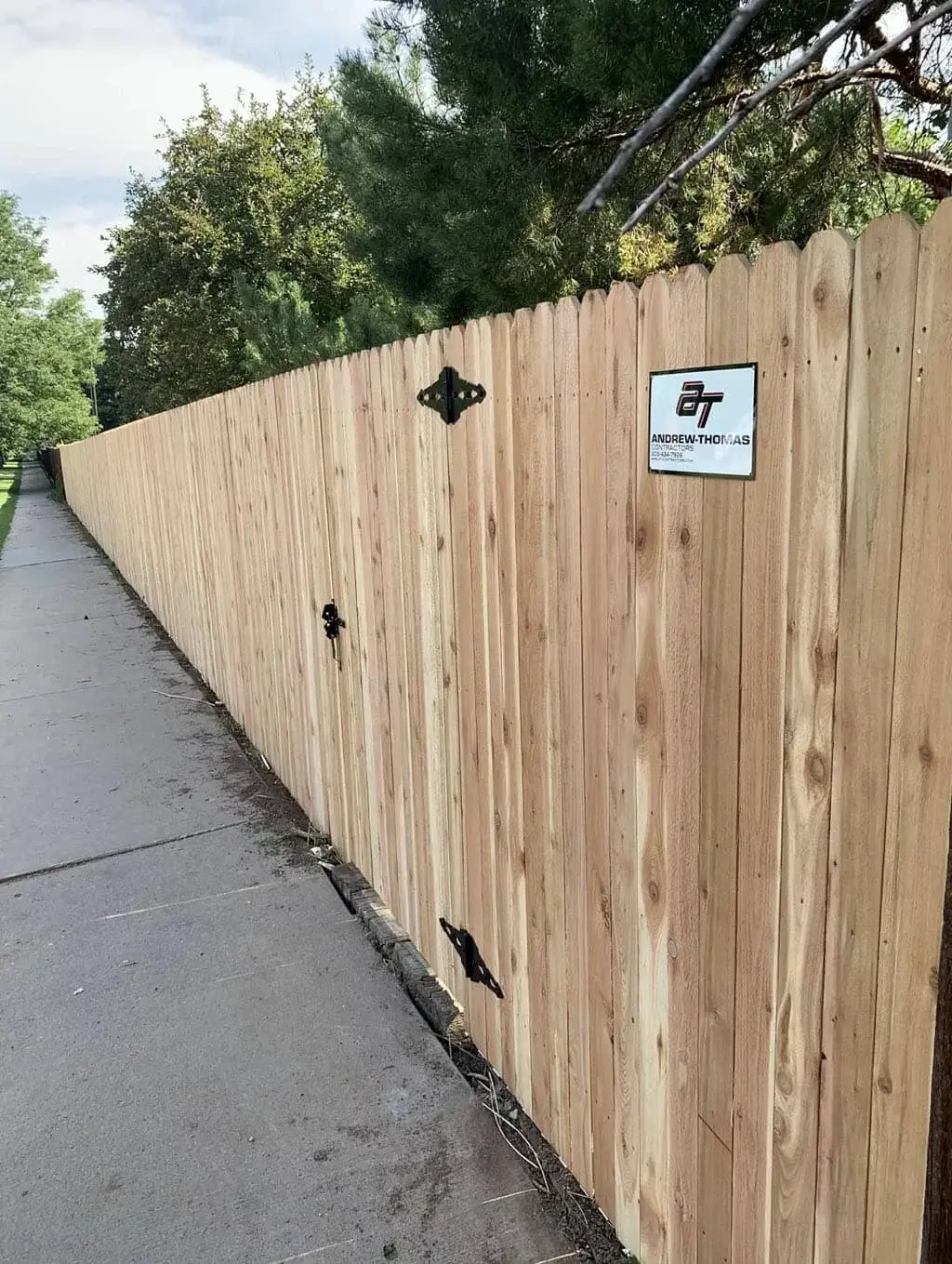 Wooden privacy fence runs along a concrete sidewalk, with a small sign attached and trees beyond it.