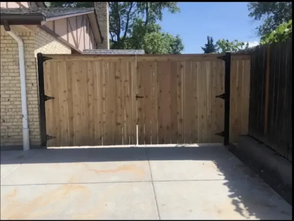 Wooden backyard gate with black metal hinges closes a concrete driveway between a brick house and a wooden fence.
