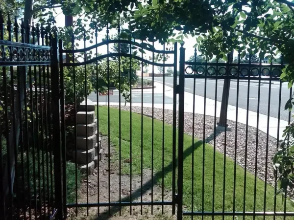 Iron decorative gate opening to a sidewalk and street, with a gravel border and green lawn on either side, and leafy shrubs above