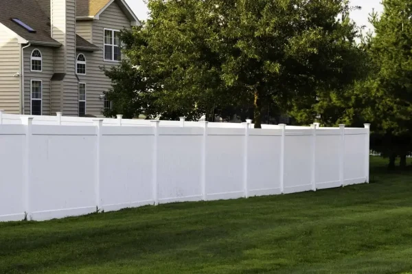 White vinyl privacy fence runs along a green lawn in a suburban backyard with a beige house and trees nearby.