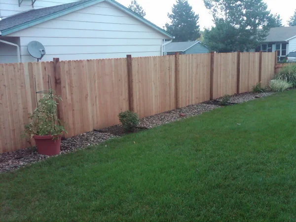Backyard view with a tall wooden privacy fence, a satellite dish on a light-colored house, and a gravel border beside the green lawn.