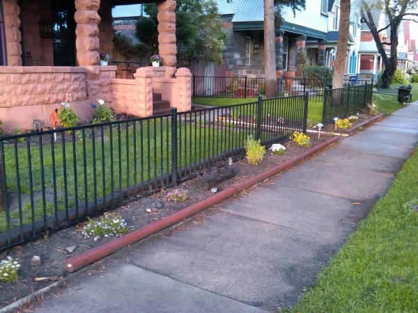 Residential sidewalk with a black metal fence and stone pillars framing a front yard with flowers; a red pipe lies along the curb edge.